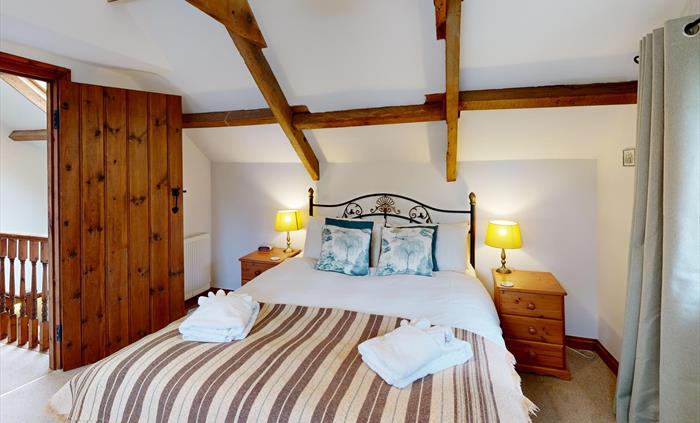 A bright bedroom with exposed wooden beams, double bed, wooden wardrobe, and soft lighting.
